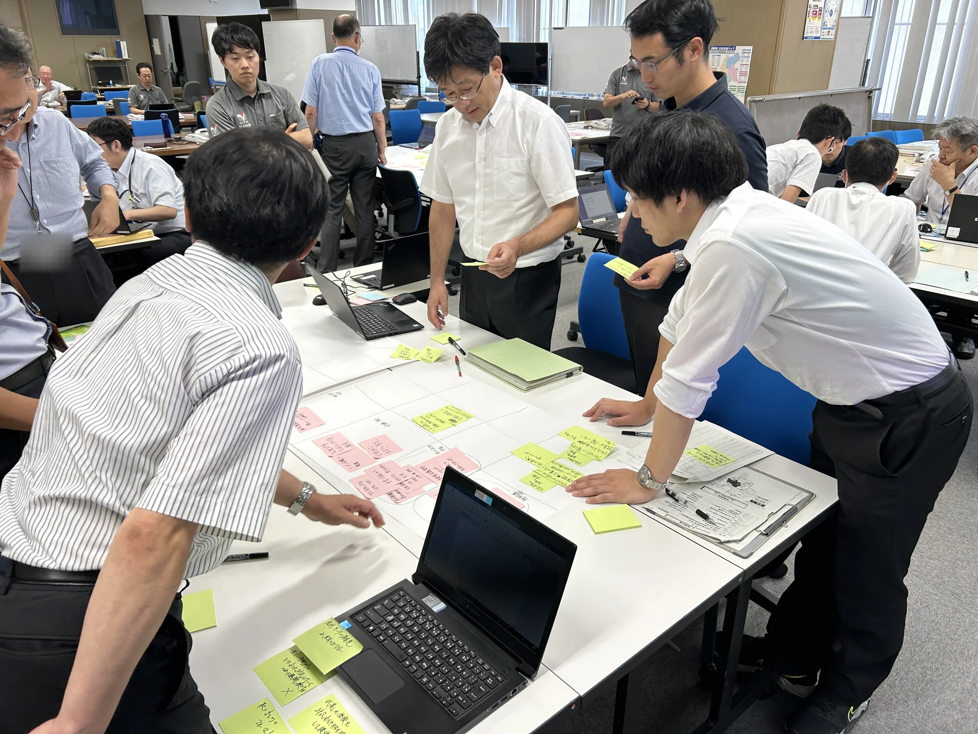 訓練のふりかえりで、参加者が机に広げた模造紙と付箋を囲んで話し合っている様子。